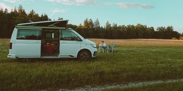 Ein Camper steht auf einer Wiese. Eine Frau sitzt daneben an einem Tisch und isst.