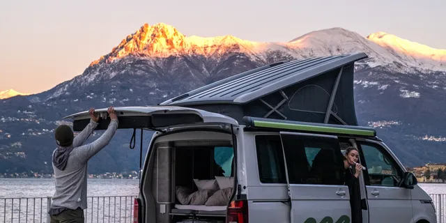 Ein junger Mann schließt gerade den Kofferraum seines Off Campers, auf dem Beifahrersitz putzt eine junge Frau Zähne. Dahinter ist ein See und ein Berg mit Schnee zu sehen.