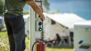 Landstrom Camper: Ein Mann schließt seinen Camper auf dem Campingplatz an den Landstrom an, im Hintergrund sieht man weiße Wohnmobile.
