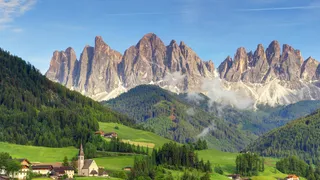 The Dolomites have many beautiful places to offer: a mountain massif in front of blue sky, in the valley you can see an idyllic place with a church