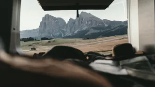 A man who explores the most beautiful places in South Tyrol with the camper, looks out of the Camper's window on the mountain landscape, next to him lies a hat