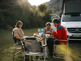 Three friends: breakfast in front of their off camper on the meadow in a mountain landscape while the sun shines.