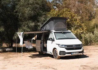 A camper is close to a forest with an extended awning.