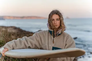 A woman with a surfboard under the arm is on the beach.