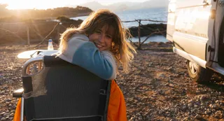 Lachende Frau auf Campingstuhl neben Off Camper beim Sonnenuntergang