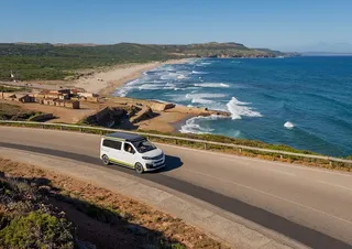Ein VW California Van fährt eine K üstenstraße entlang. Im Hintergrund erstreckt sich das blaue Meer.
