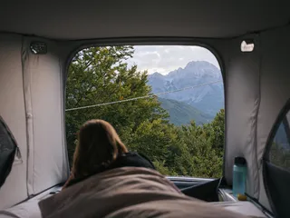Eine Frau liegt in einem Camper im Bett und blickt auf eine bewaldete Berglandschaft.
